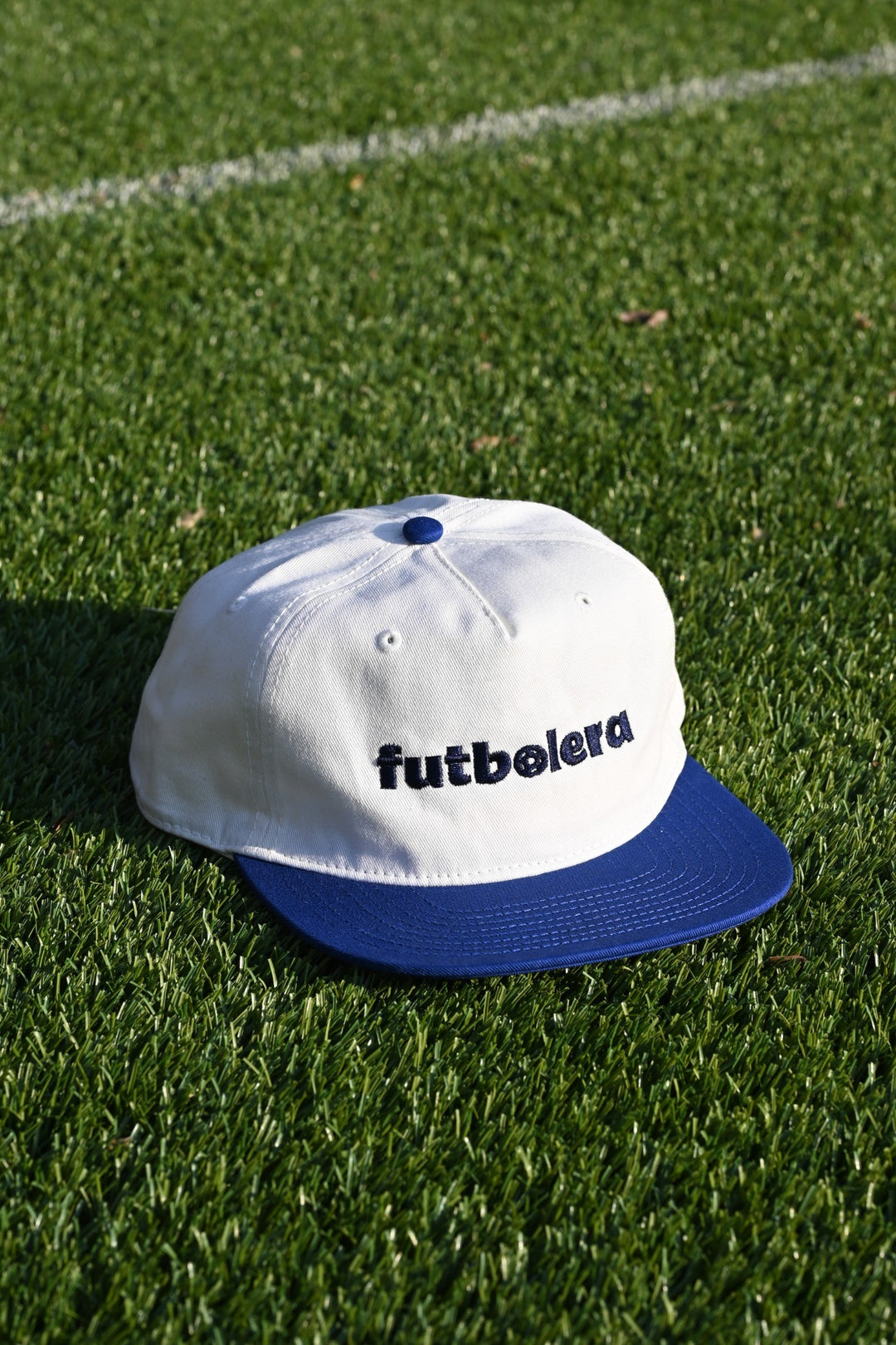 White cap with blue brim and 'futbolera' logo on grass