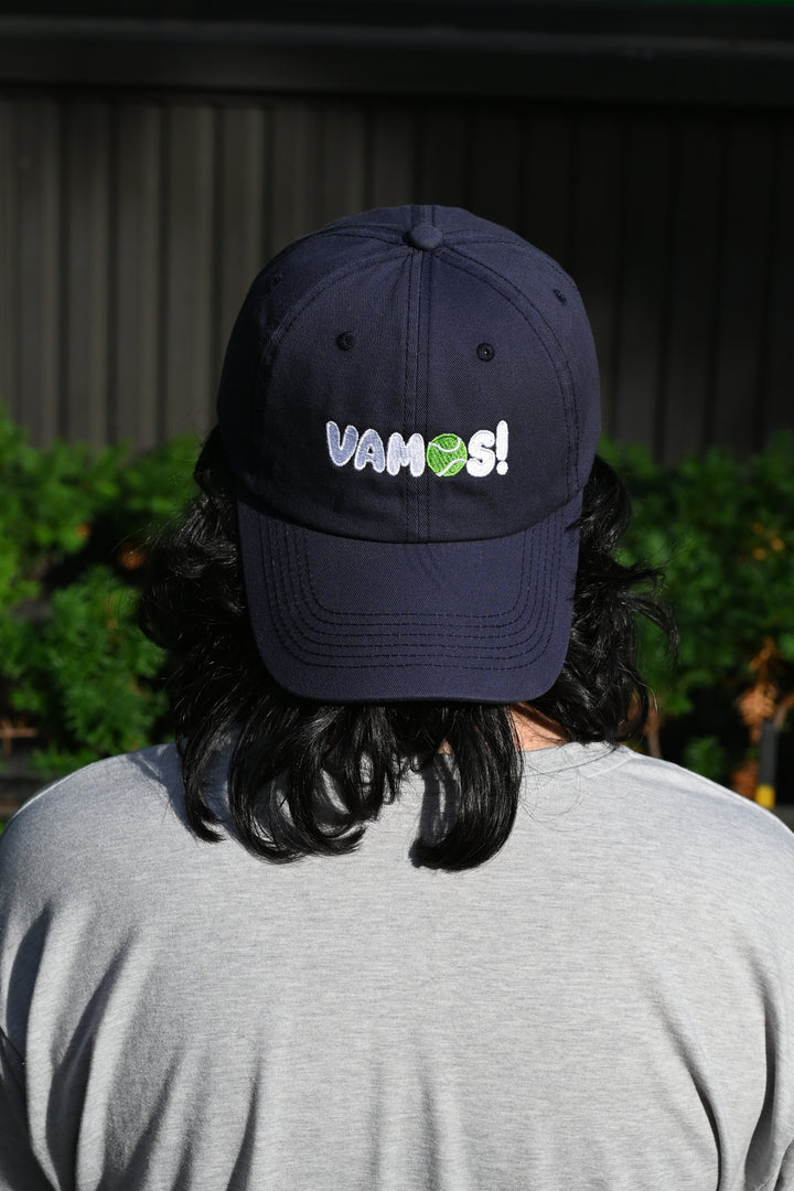 A person viewed from behind wearing a dark navy tennis cap with the text &