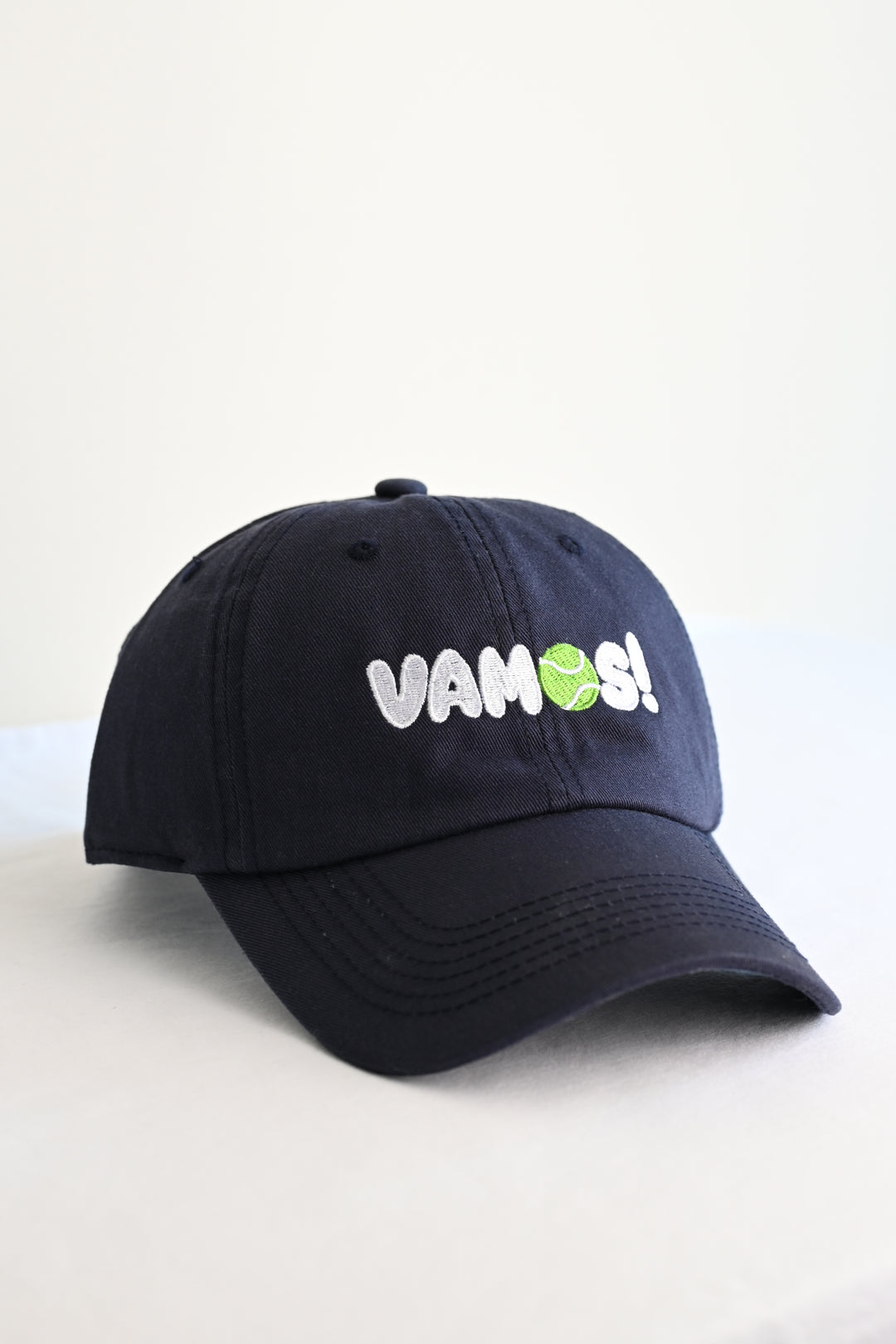 A navy blue tennis cap with 'VAMOS!' embroidered on the front in white and green, set against a plain off-white background.