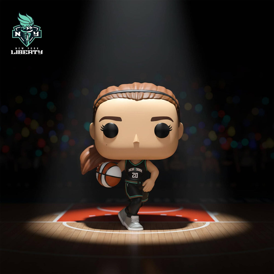 A stylized basketball player Sabrina Ionescu collectible figure stands on a basketball court under a spotlight, holding a basketball. The figure has a peach face and skin tone, a teal hair headband, a black jersey with teal accents and the number 20, and white sneakers.